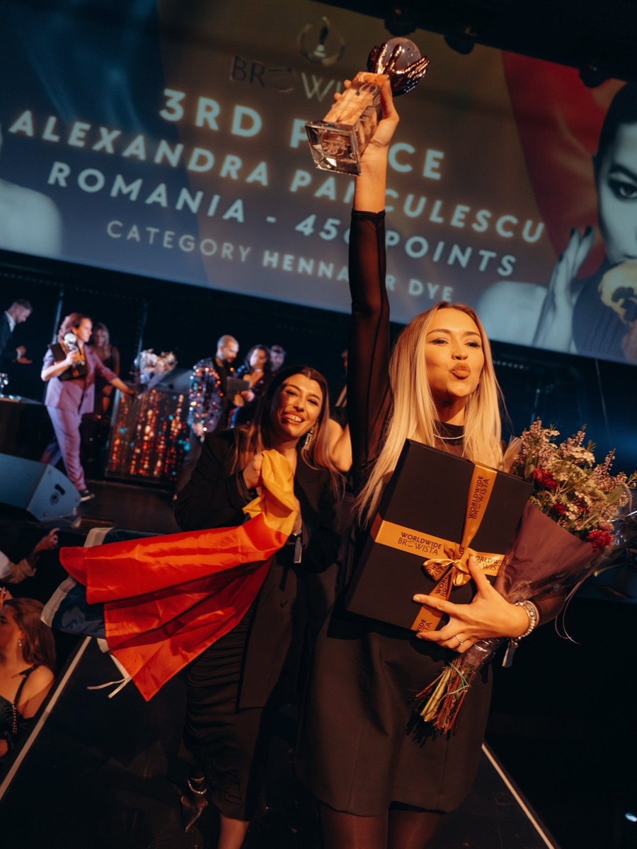 Concurentă din România pe scena din Rotterdam, cu trofeul de cristal ridicat, iar în spatele ei o colegă poartă tricolorul pe umeri; pe ecran scrie locul 3, România, 450 de puncte
