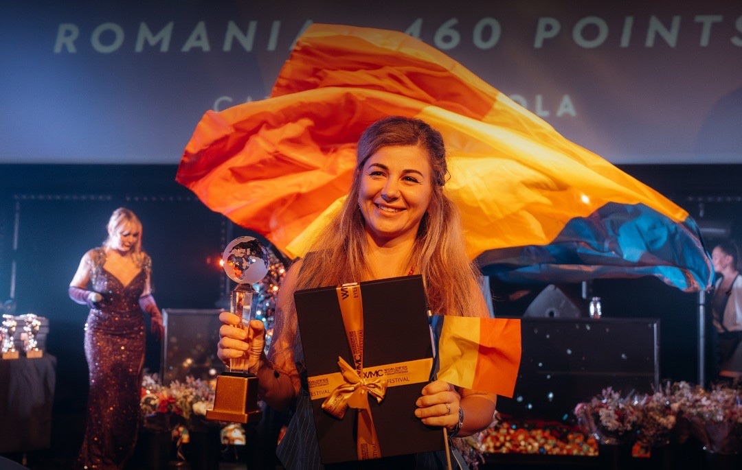Concurentă din România, zâmbind pe scenă, cu trofeul-glob, cutia de premiere și un drapel mic al României; deasupra ei flutură un tricolor mare