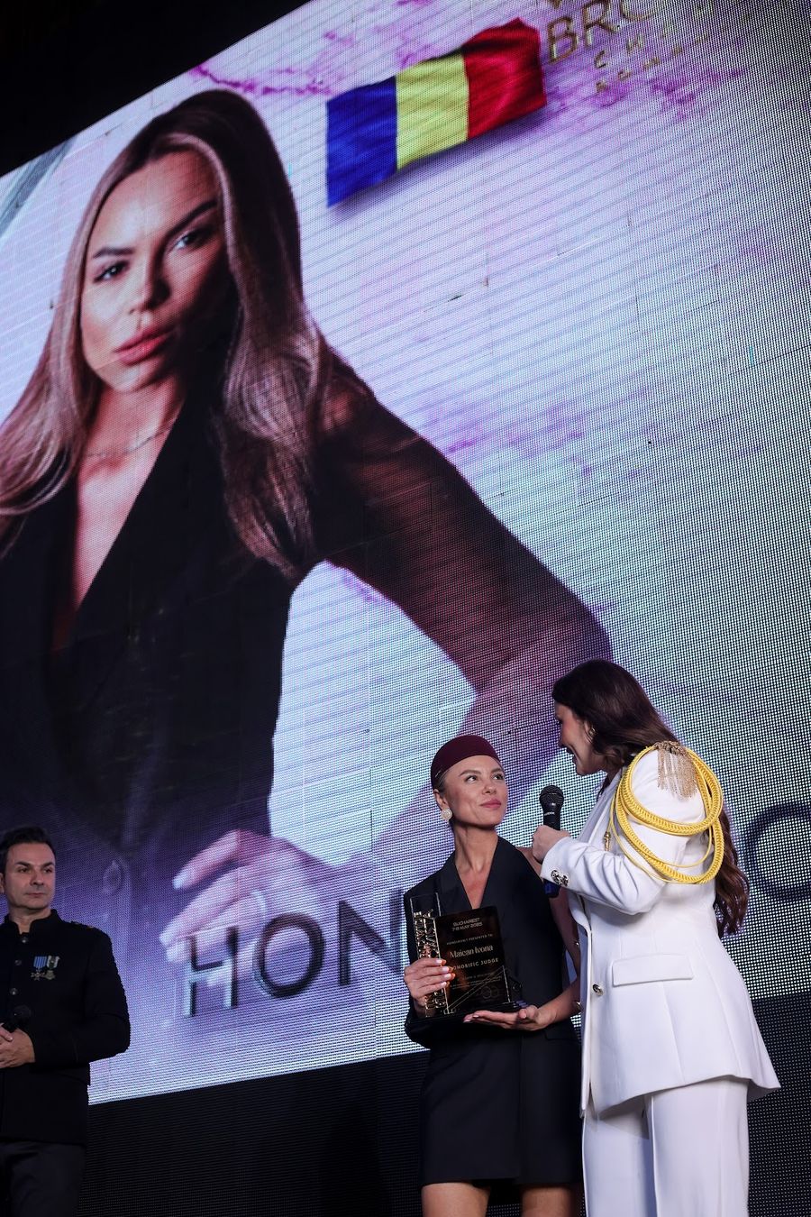 Pe scenă, o artistă cu trofeul în mână și prezentatoarea; în spate, portretul ei pe ecranul mare, cu tricolorul în colț