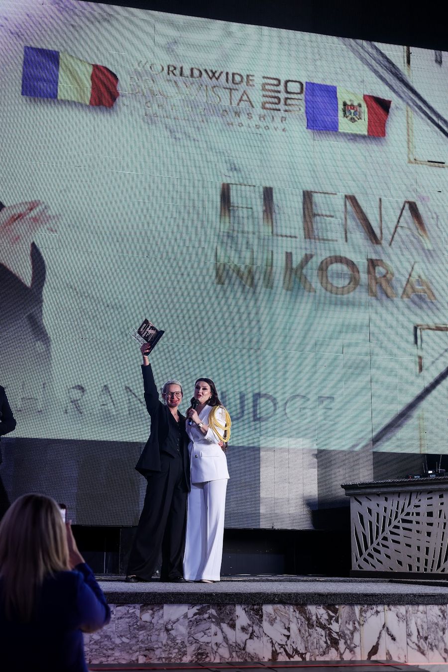Două persoane pe scenă, una cu trofeul ridicat, sub steagurile României și Moldovei de pe ecran